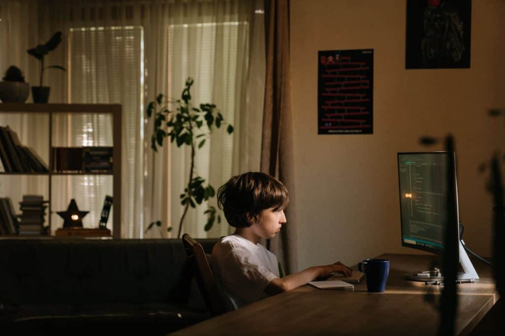 A young boy sitting at a wooden desk at night intently looking at a computer screen filled with lines of code.