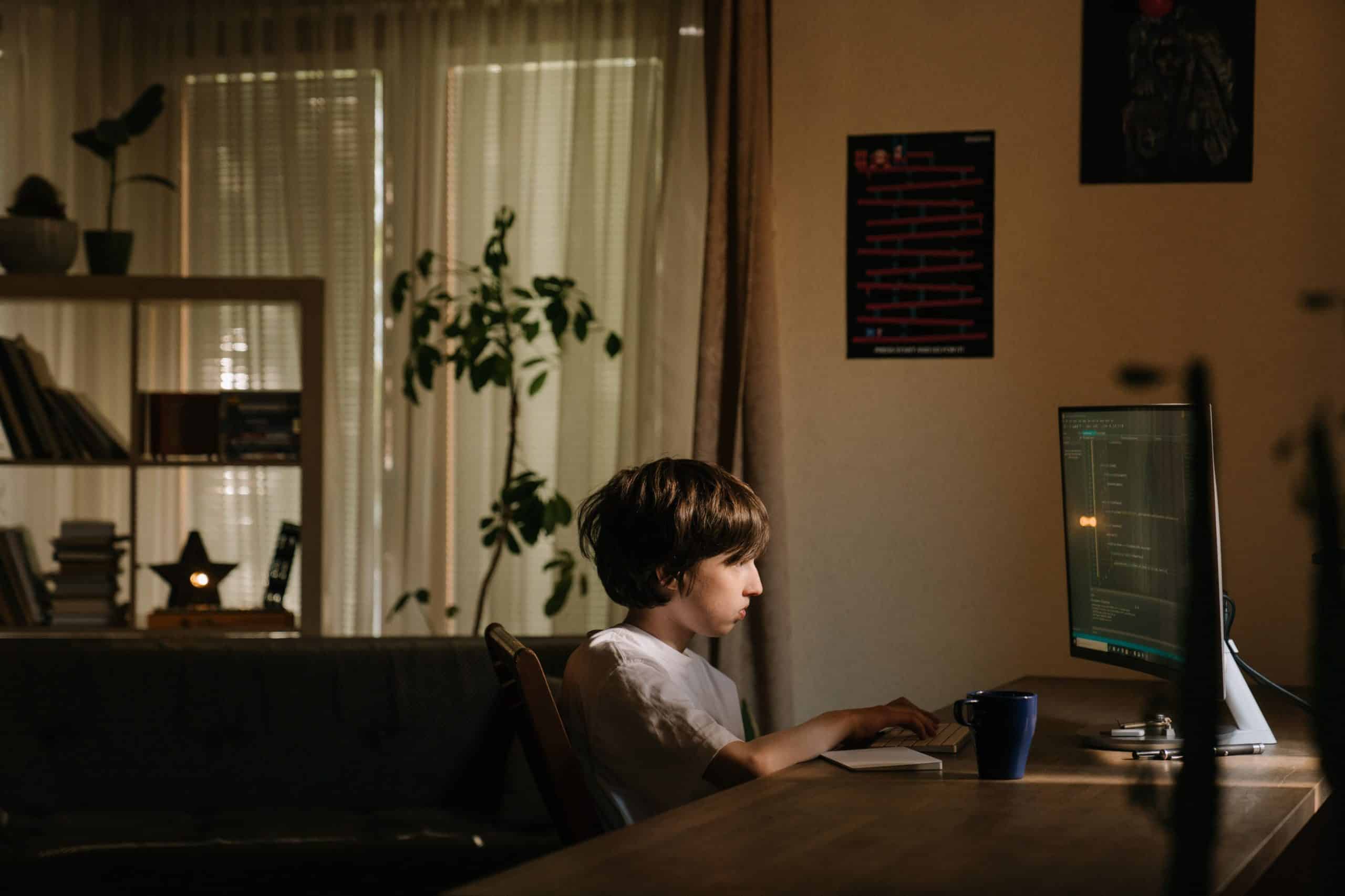 A young boy sitting at a wooden desk at night intently looking at a computer screen filled with lines of code.